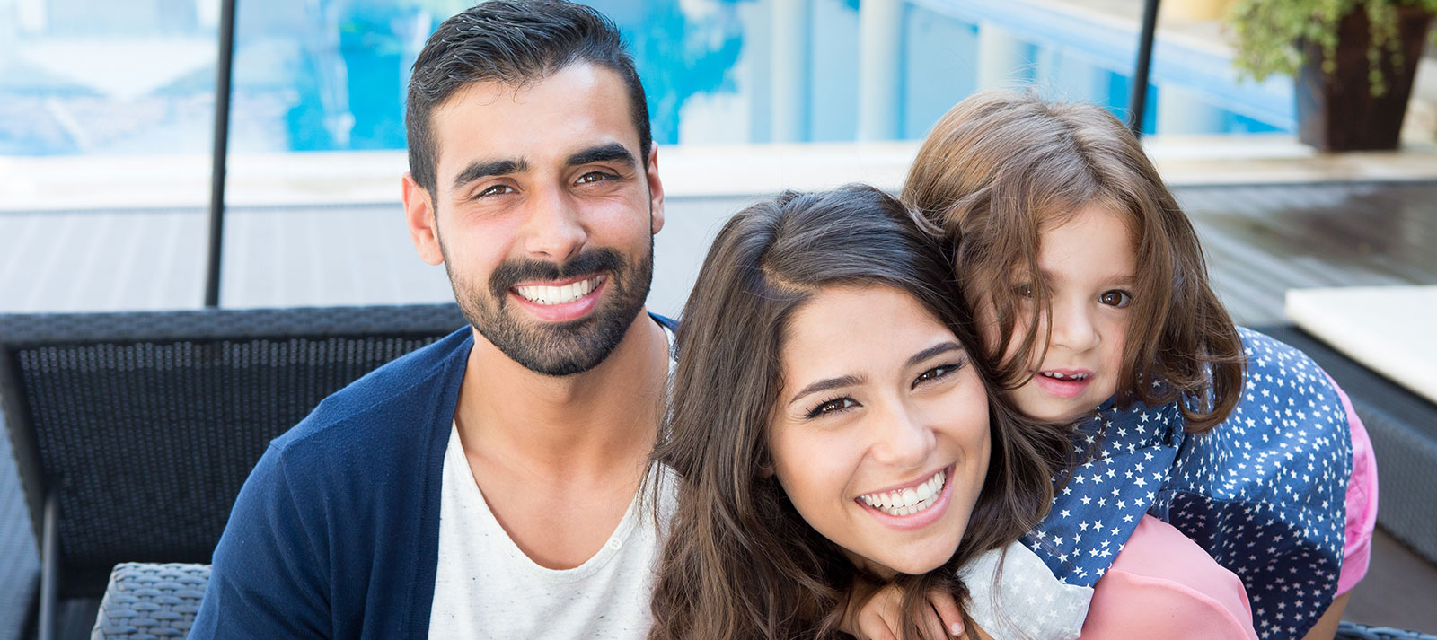 A family of four poses together for a photo at an outdoor dining area with a man wearing a blue sweater, a woman in a white shirt, and two children smiling at the camera.
