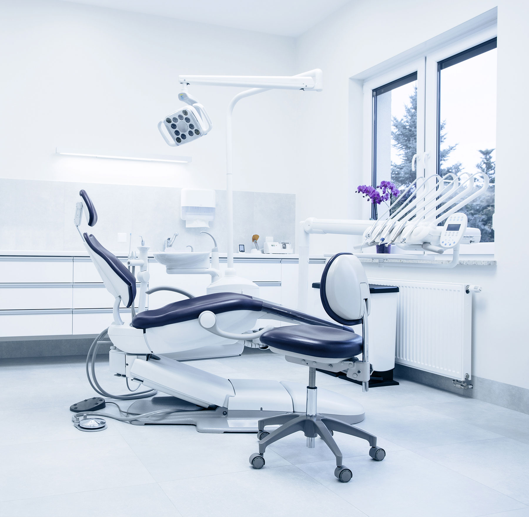 The image shows a modern dental office interior with a large dental chair in the foreground, a dental examination table, a dental equipment cabinet, a sink, and a window letting in natural light.